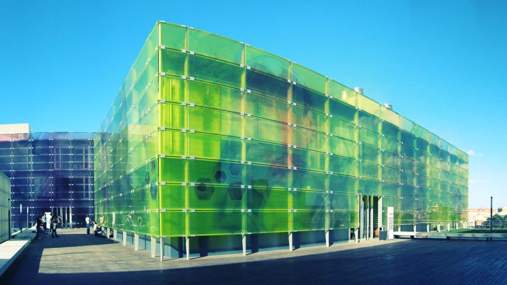 Edificio de fachada de vidrio verde y azul de la Ciudad Politécnica de la Innovación, en el campus de Vera de Valencia, bajo un cielo despejado.