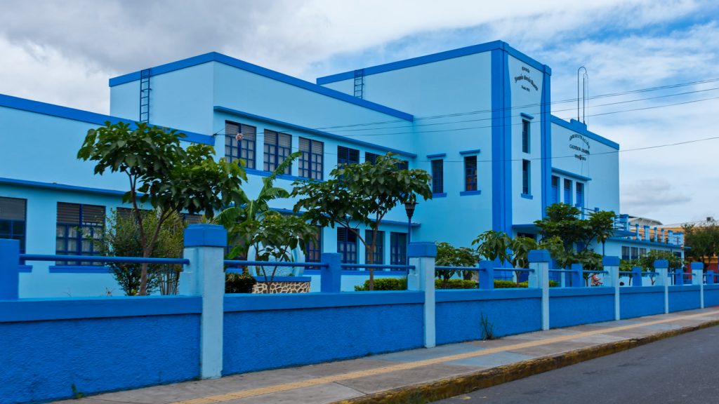 Fachada de la Escuela Joaquín García Monge, un edificio escolar pintado de azul claro y azul intenso, con verja y árboles, en Desamparados, Costa Rica.