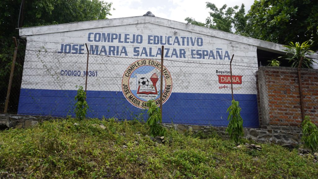 Muro encalado de un centro educativo salvadoreño con el rótulo pintado «Complejo Educativo José María Salazar España» y el escudo del centro, rodeado de vegetación.