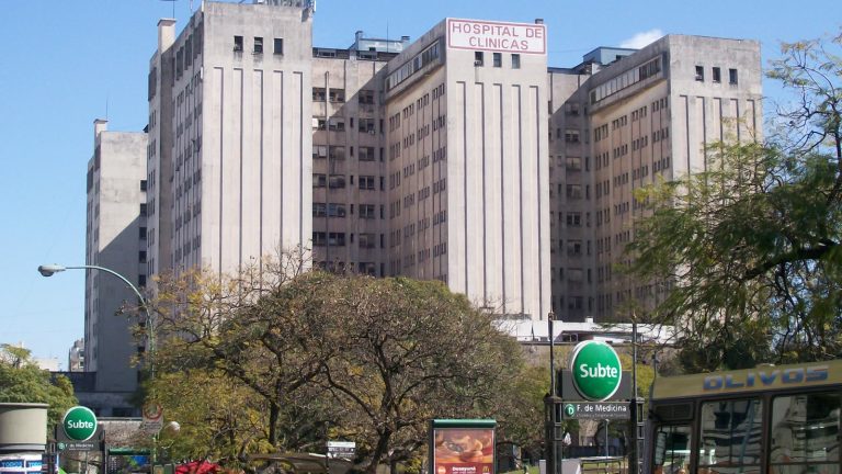 Fachada del Hospital de Clínicas José de San Martín, en Buenos Aires: un gran edificio racionalista de hormigón con el cartel rojo del hospital en lo alto y árboles y una entrada de subte en primer plano.