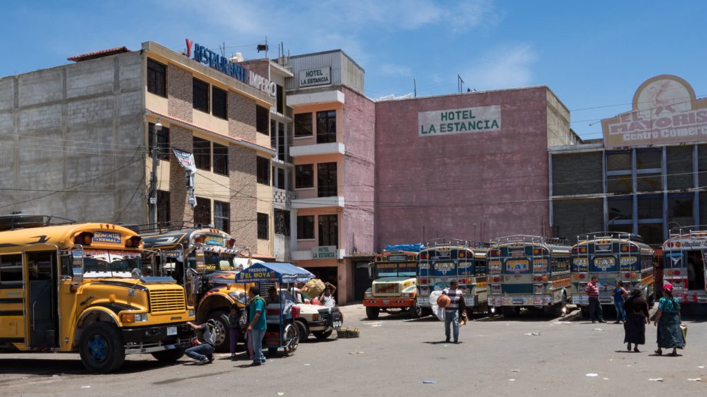 Terminal de autobuses de Huehuetenango, en Guatemala: filas de buses escolares pintados de colores, vendedores ambulantes y viajeros cruzando la explanada bajo un cielo despejado.