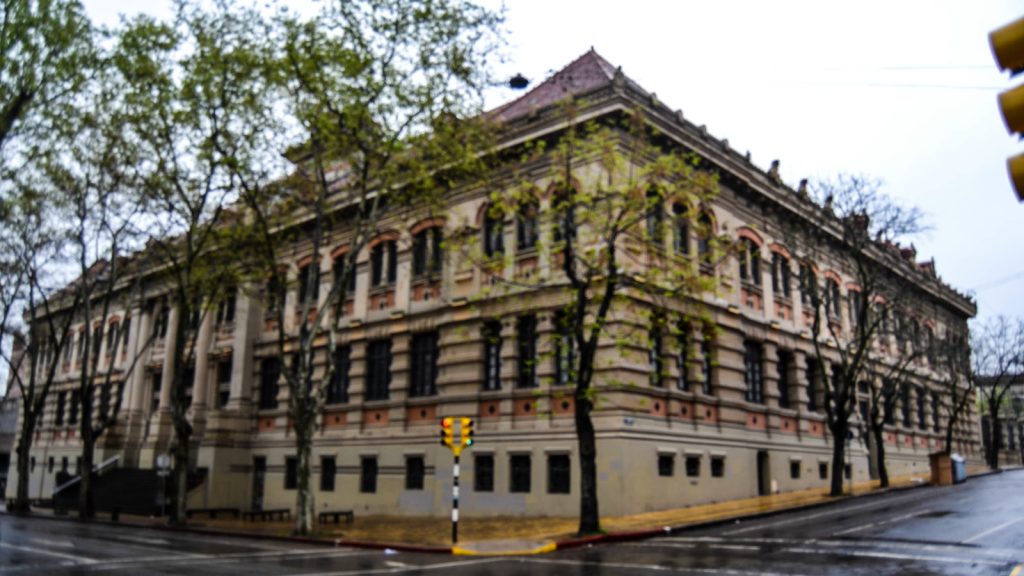 Fachada del Instituto Alfredo Vásquez Acevedo, un edificio histórico de tres plantas con arcos y ladrillo a la vista, en una esquina arbolada de Montevideo en un día de lluvia.
