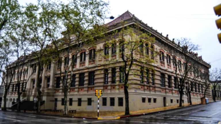 Fachada del Instituto Alfredo Vásquez Acevedo, un edificio histórico de tres plantas con arcos y ladrillo a la vista, en una esquina arbolada de Montevideo en un día de lluvia.