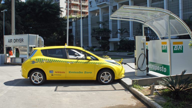 Taxi eléctrico amarillo Nissan Leaf con el rótulo «Emissão Zero» conectado a un punto de carga bajo una marquesina en una estación de servicio de Río de Janeiro, con edificios de viviendas al fondo.