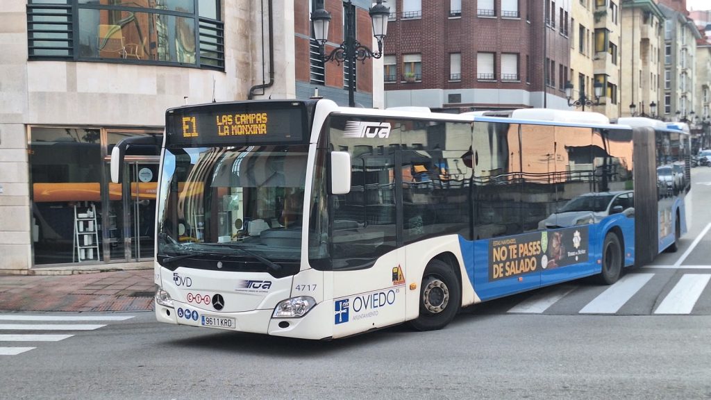 Autobús urbano articulado blanco y azul de la línea E1 de TUA, con los logotipos de Oviedo y del CTA, girando en un paso de cebra de una calle de Oviedo.