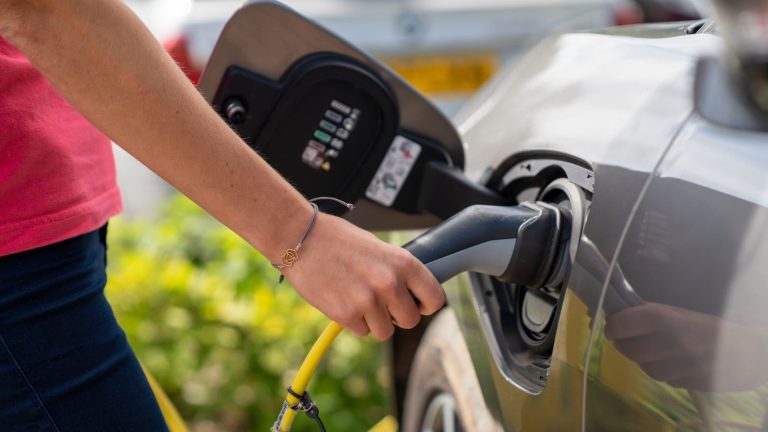 Primer plano de una mano que conecta el cargador de un punto de recarga a un coche eléctrico gris, con un cable amarillo y vegetación al fondo.