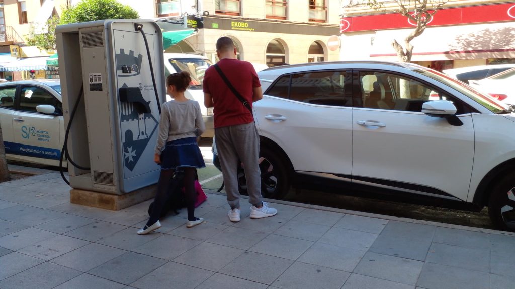 Un hombre y una niña de espaldas junto a un punto de recarga municipal en una acera de Amposta, con un todoterreno eléctrico blanco aparcado y conectado al lado.